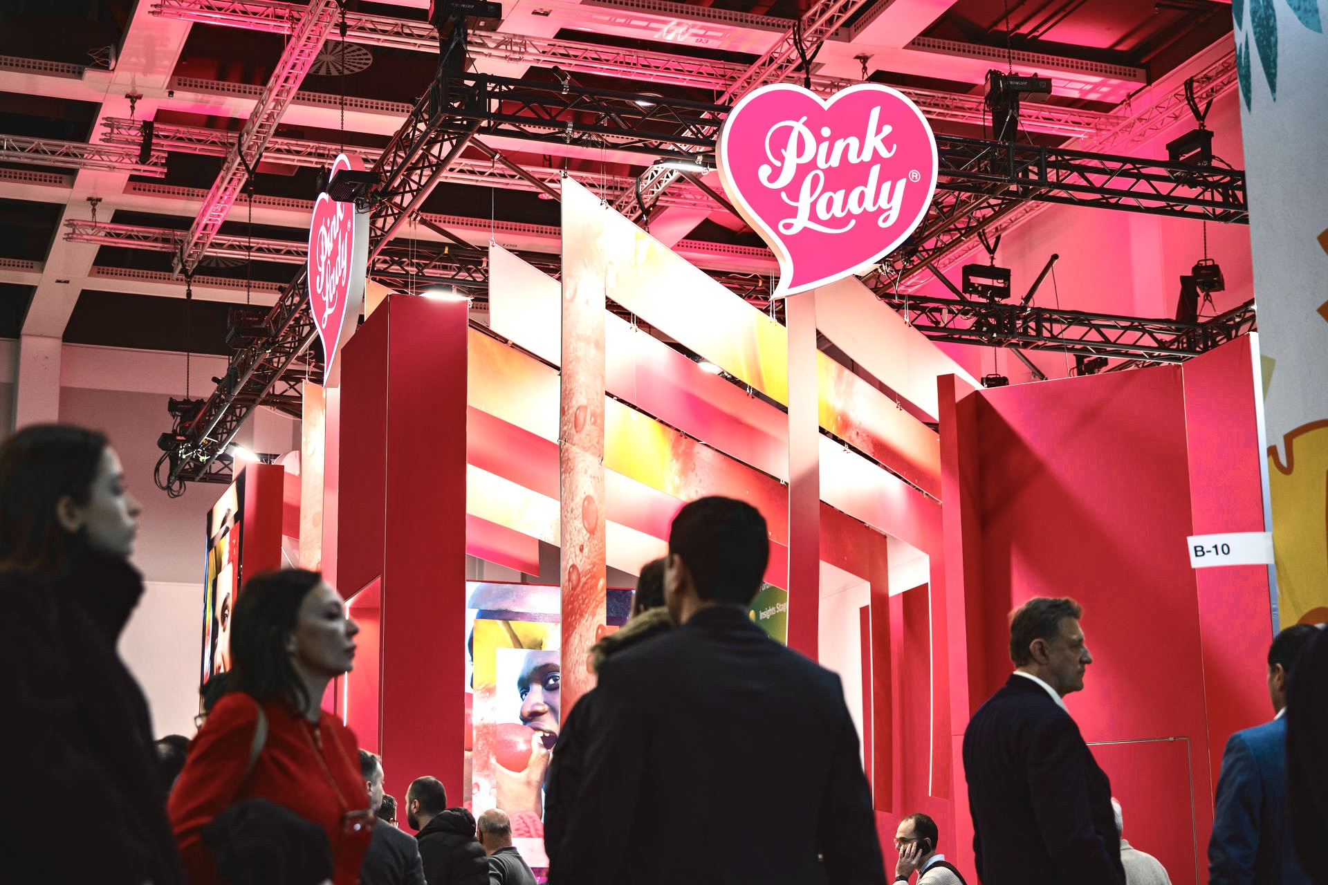 Stand Pink Lady Europe Fruit Logistica Berlin en activité — vue depuis l'allée avec visiteurs, structure colonnes rouge et rose, scénographie événementielle WIO